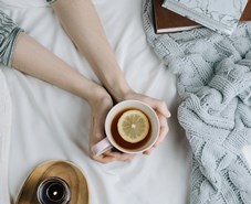 Cozy flatlay of woman's hand holding lemon tea in bed with white sheets and blanket, knitted sweater and candle